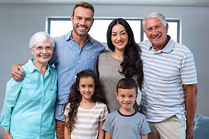 A family of five, including an elderly woman, a man, a woman, and two children, are standing and smiling for a photo in a room with a gray wall and a window.