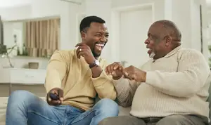 Two men sitting on a couch, one wearing a beige sweater and the other wearing a yellow sweater, both smiling and holding a remote, with a white cabinet and a plant on the left, and a white door behind them
