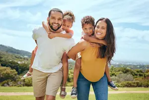 A family of three, including a man, a woman, and a child, are standing in a grassy field, smiling and posing for a photo with the man holding the child on his shoulders. They are surrounded by lush greenery, and in the distance, there are mountains under a clear blue sky with scattered clouds.