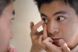 A person is seen in a mirror, inserting an eye contact lens, with another person standing nearby observing, both in a room with white walls
