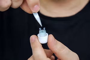 A person is holding a small white object with a transparent cover and a silver tip in their fingers, probably putting a drop of medicine on it.