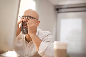A bald man with a beard and glasses is sitting in front of a computer monitor with his hands on his face.