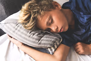 A boy sleeps on his side on a bed with a pillow supporting his head and neck.