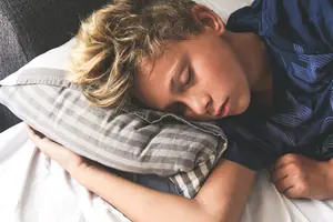 A boy sleeps on his side on a bed with a pillow supporting his head and neck.