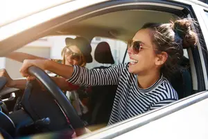 Two women sitting inside a white car and smiling while one of them is driving and the other is holding the steering wheel