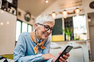A smiling woman with glasses and a scarf using a tablet in a room with a counter and plants