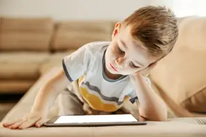A young boy with his eyes closed is sitting on a couch while looking at a tablet.