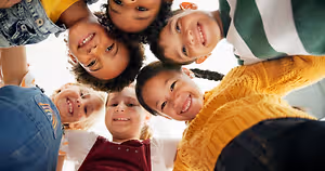A group of children smiling and standing in a circle with one girl wearing a yellow sweater and black pants in the center