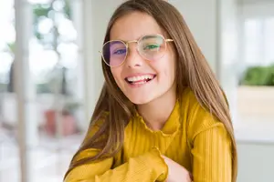 Close-up of a smiling girl wearing sunglasses and a yellow shirt in a room with a glass window in the background.