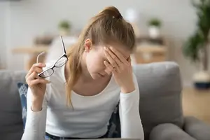 A woman holding glasses with a hand on her forehead in a living room.