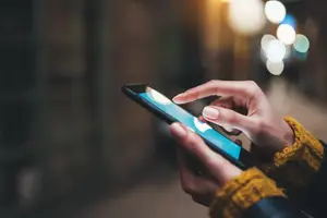 A person wearing a yellow sweater is holding a smartphone with a blue screen in front of a blurry background with a few lights.