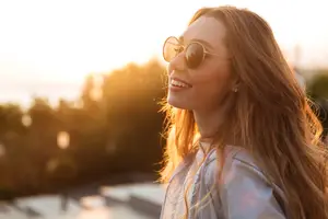 Portrait of a smiling woman with long hair wearing sunglasses and a gray jacket standing in an outdoor area during sunset.