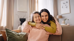 Two women embracing on a sofa, one wearing glasses and the other in a yellow long-sleeve shirt, with a picture frame and lamp in the background.