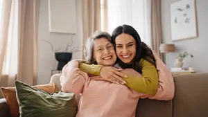 Two women embracing on a sofa, one wearing glasses and the other in a yellow long-sleeve shirt, with a picture frame and lamp in the background.