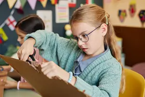 A girl wearing glasses and a sweater is cutting a piece of cardboard in a classroom.