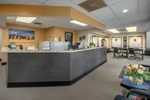 An empty reception area with a desk, a monitor, and a flower vase, along with several chairs, a table, and a poster on the wall.