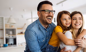 A man and a woman with their young daughter are smiling and hugging each other inside a kitchen