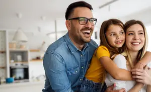 A man and a woman with their young daughter are smiling and hugging each other inside a kitchen