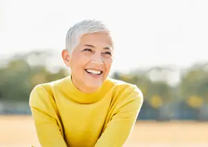 A woman with white hair smiles and poses for a picture in a yellow turtleneck shirt in an outdoor setting.
