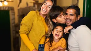 A family with two children, a woman in a yellow sweater, a man in a black sweater, and two children in yellow shirts are smiling and hugging each other in a room.