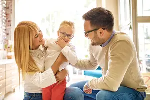 A man and woman hold a young child with glasses and they are all smiling.