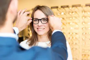 A man is adjusting a woman's glasses in a blurry background with lights and glasses frames