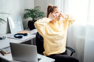 A woman is sitting in a chair in front of a desk, looking exhausted with her hands on her face.