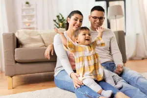 A portland or family of three is sitting on the floor and smiling for a photo in the living room.