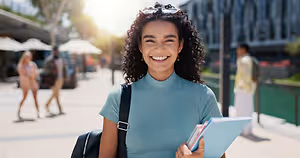 A smiling woman in a blue turtleneck holding a book and a folder while walking on a city street.
