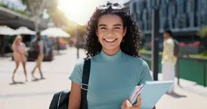 A smiling woman in a blue turtleneck holding a book and a folder while walking on a city street.