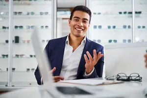 A man in a suit and white shirt smiles and gestures with his hands while sitting at a desk with a laptop and papers in front of him.