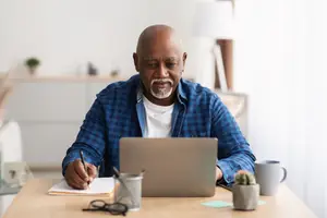 A man is sitting at a desk with a laptop, pen, notebook, and mug.
