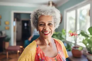 A smiling elderly woman with gray hair in a yellow shirt is standing in a room with a window and potted plants.