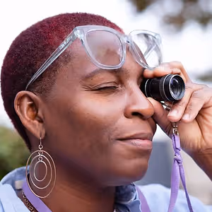 A woman with maroon hair is looking through a telescope with her eyes closed, wearing glasses and earrings