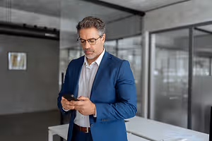A man in a blue suit is looking at his phone in an office.