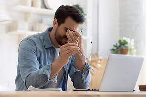 A man with glasses in his hand is sitting in front of a laptop with a blurry background.
