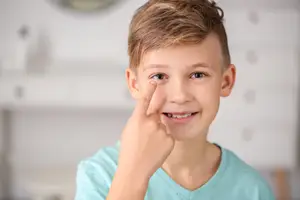 A boy wearing a blue shirt is smiling and pointing to his eye, likely in a room with white walls, possibly for an eye examination.