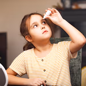 A young girl sits at a table, holding a white bottle and applying eye drops.