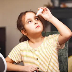 A young girl sits at a table, holding a white bottle and applying eye drops.