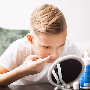 A young boy is examining his nose in a mirror while holding a small bottle of eye drops on the table beside him.
