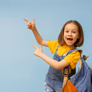 A young girl in a yellow shirt and denim overalls is pointing with her right hand and smiling while carrying a blue backpack with white dots, possibly posing for a photo against a blue background.