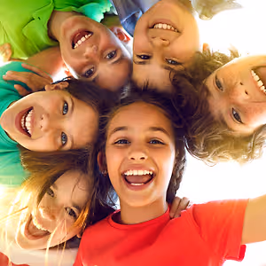 A group of smiling children in a circle wearing green and red shirts, with sunlight shining from above.