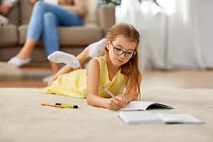 A young girl in a yellow dress is sitting on the carpet, doing her homework with a pencil and books on the floor while a woman is sitting on the couch behind her.