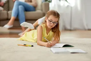 A young girl in a yellow dress is sitting on the carpet, doing her homework with a pencil and books on the floor while a woman is sitting on the couch behind her.