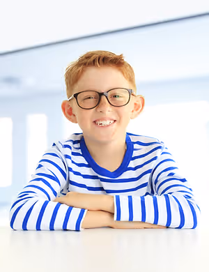 A smiling boy with glasses and a striped shirt sitting on a table.