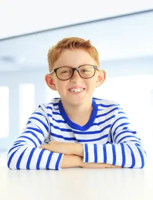 A smiling boy with glasses and a striped shirt sitting on a table.