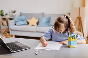 A young girl wearing glasses and a blue blouse is seated at a desk, writing on a piece of paper. A laptop and a pencil stand are on the desk. Behind her, a couch with pillows and a lamp is visible in the background.