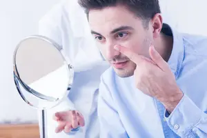 A man touches his nose with his index finger in front of a mirror, wearing a light blue shirt and another person in a white shirt behind him.
