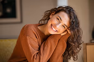 A smiling woman with curly hair sitting on a couch with a wooden table behind her.