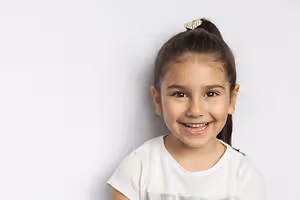 A smiling little girl with her hair tied in a ponytail is standing in front of a white wall wearing a white t-shirt with a silver design